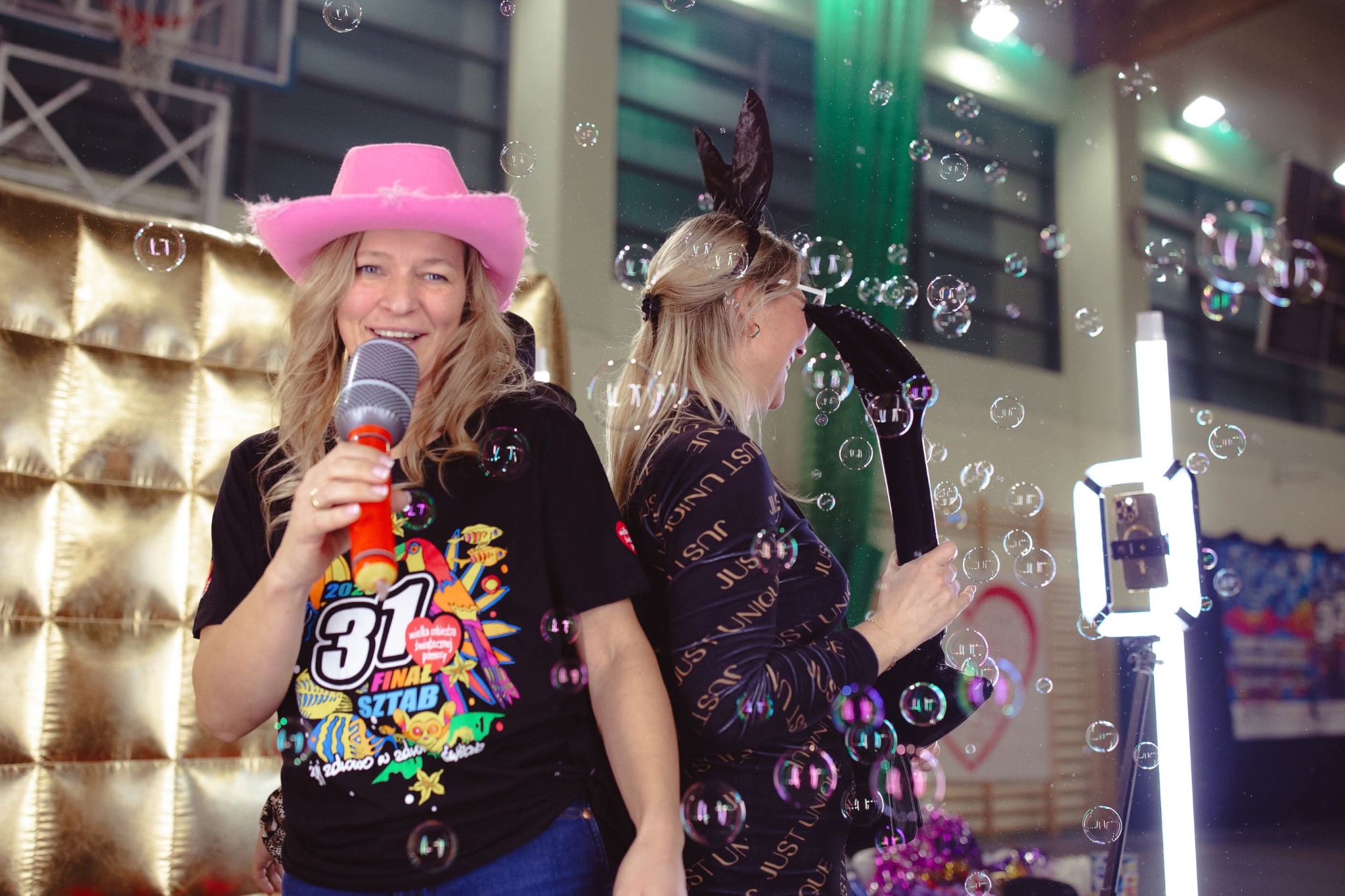 Dwie kobiety w trakcie imprezy: jedna w różowym kapeluszu śpiewa do mikrofonu, druga w opasce z uszami królika puszcza bańki mydlane z urządzenia. W tle widoczna ściana dekoracyjna i oświetlenie.