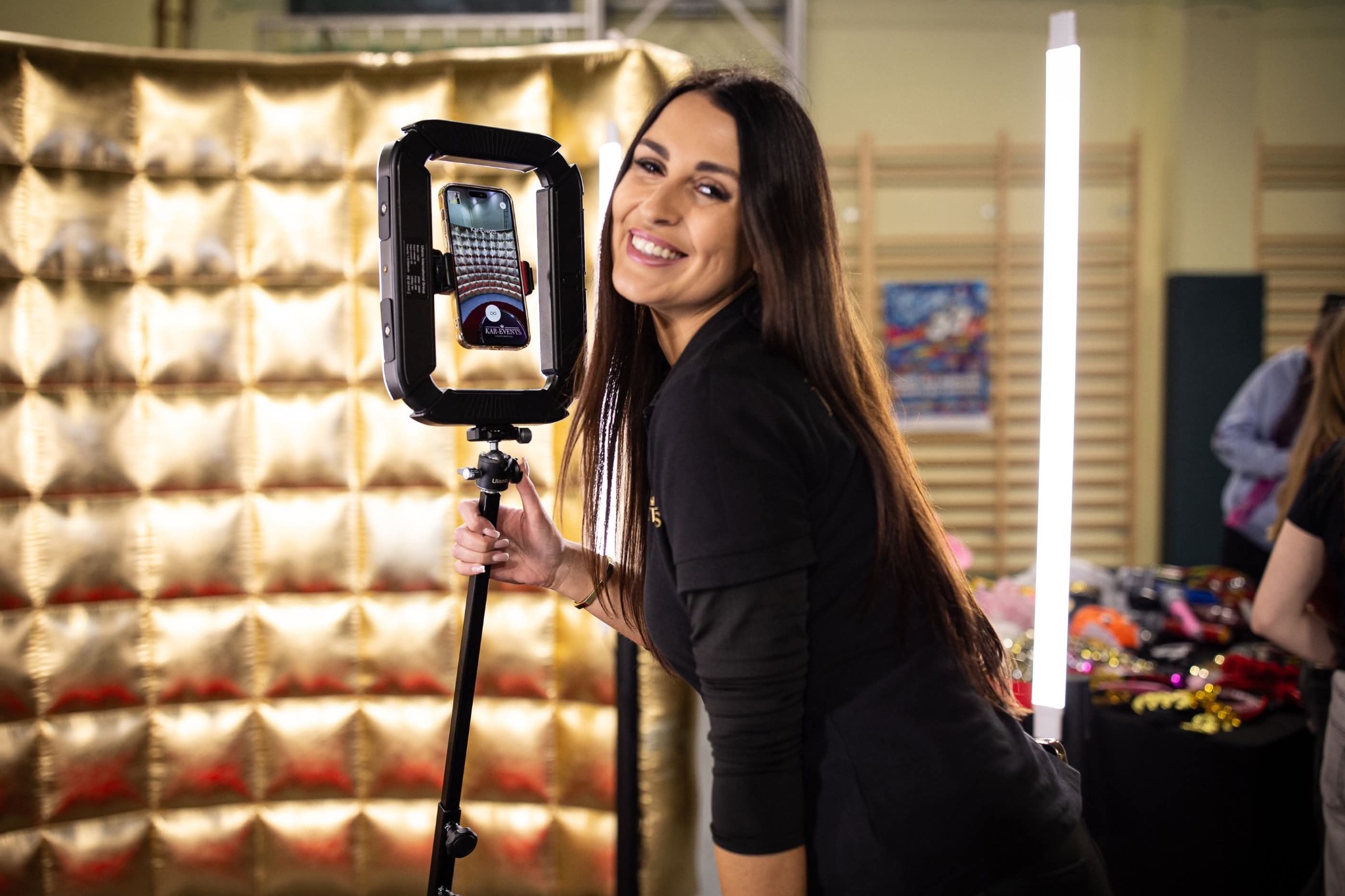 Uśmiechnięta brunetka w czarnej bluzce pozuje obok fotobudki z widocznym ekranem smartfona, w tle złota, pikowana ścianka i rekwizyty na stole.