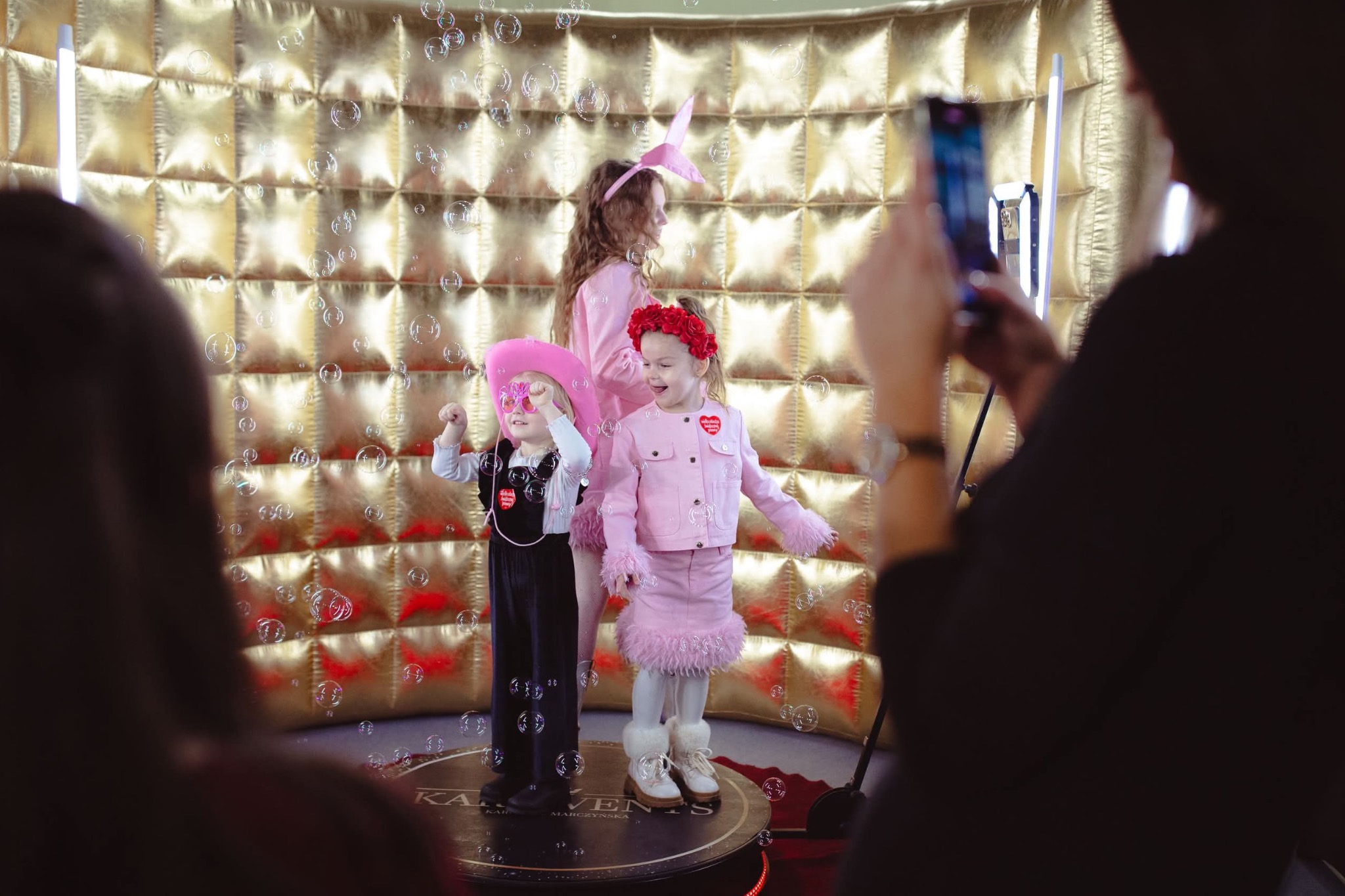 Trzy dziewczynki w przebraniach pozują w złotej fotobudce, otoczone bańkami mydlanymi, fotografowane telefonem.