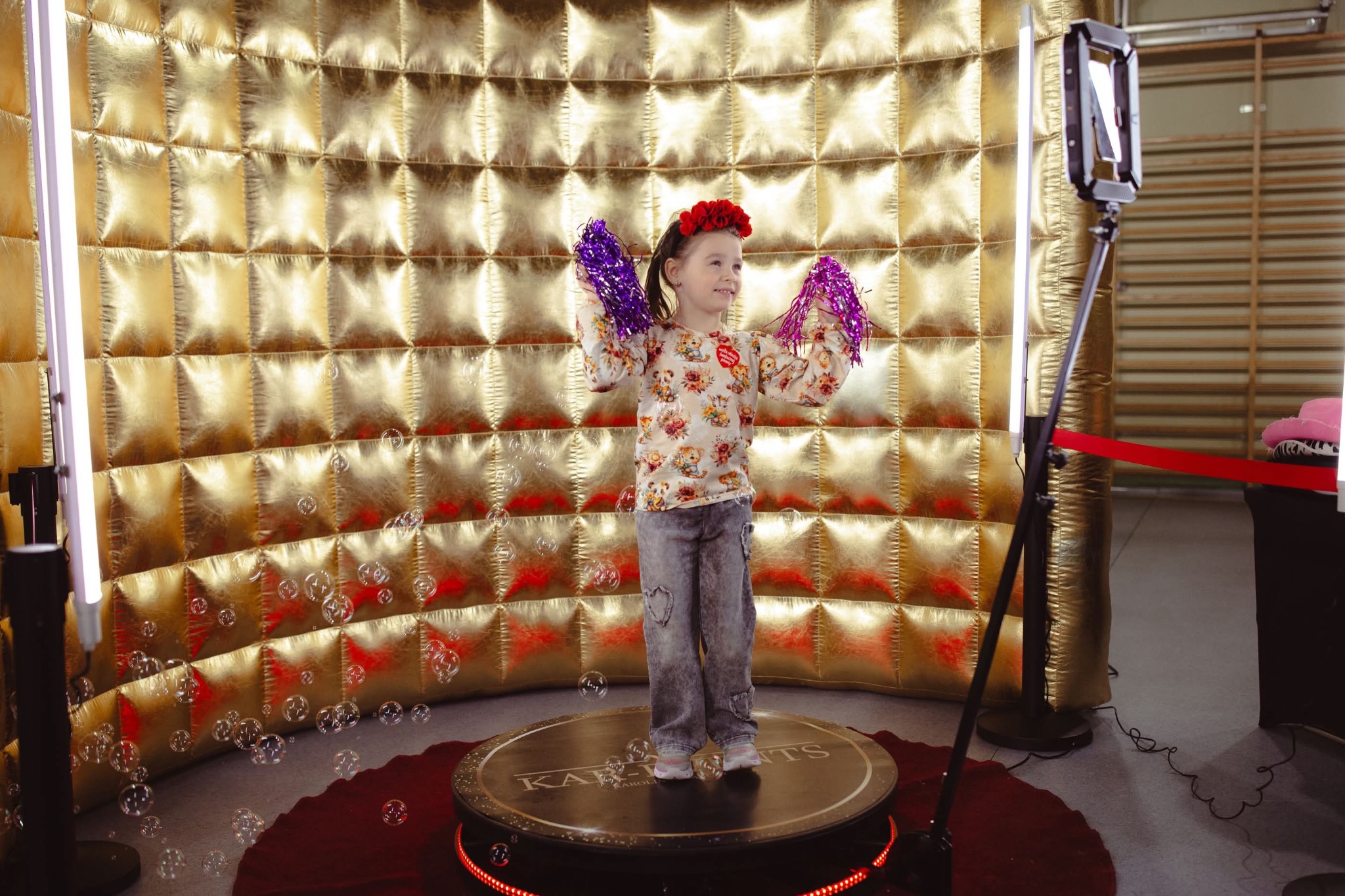 Uśmiechnięta dziewczynka z czerwonym wiankiem na głowie i fioletowymi pomponami w rękach pozuje na obrotowej platformie w złotej, dmuchanej fotobudce, otoczonej bańkami mydlanymi.