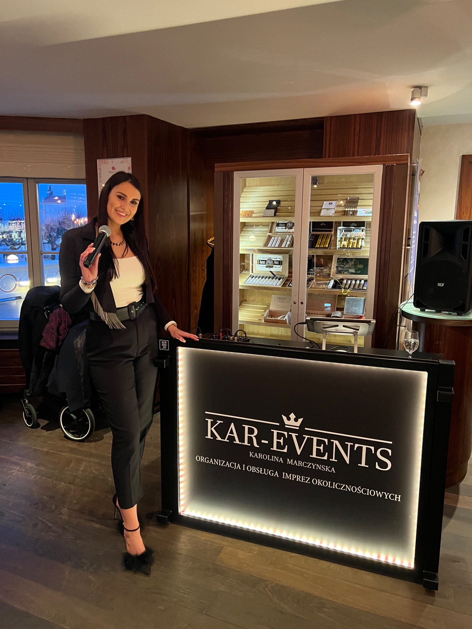 Uśmiechnięta kobieta w czarnym garniturze i mikrofonem, stojąca za podświetlanym panelem z logo 'KAR-EVENTS', obok wózka dziecięcego i głośnika RCF.