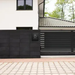 Nowoczesna, pozioma brama wjazdowa z czarnych profili w Nowej Soli, przylegająca do ściany z ciemnej, kwadratowej cegły z logo firmy. Widoczny fragment domu z białą elewacją i ciemnymi oknami.