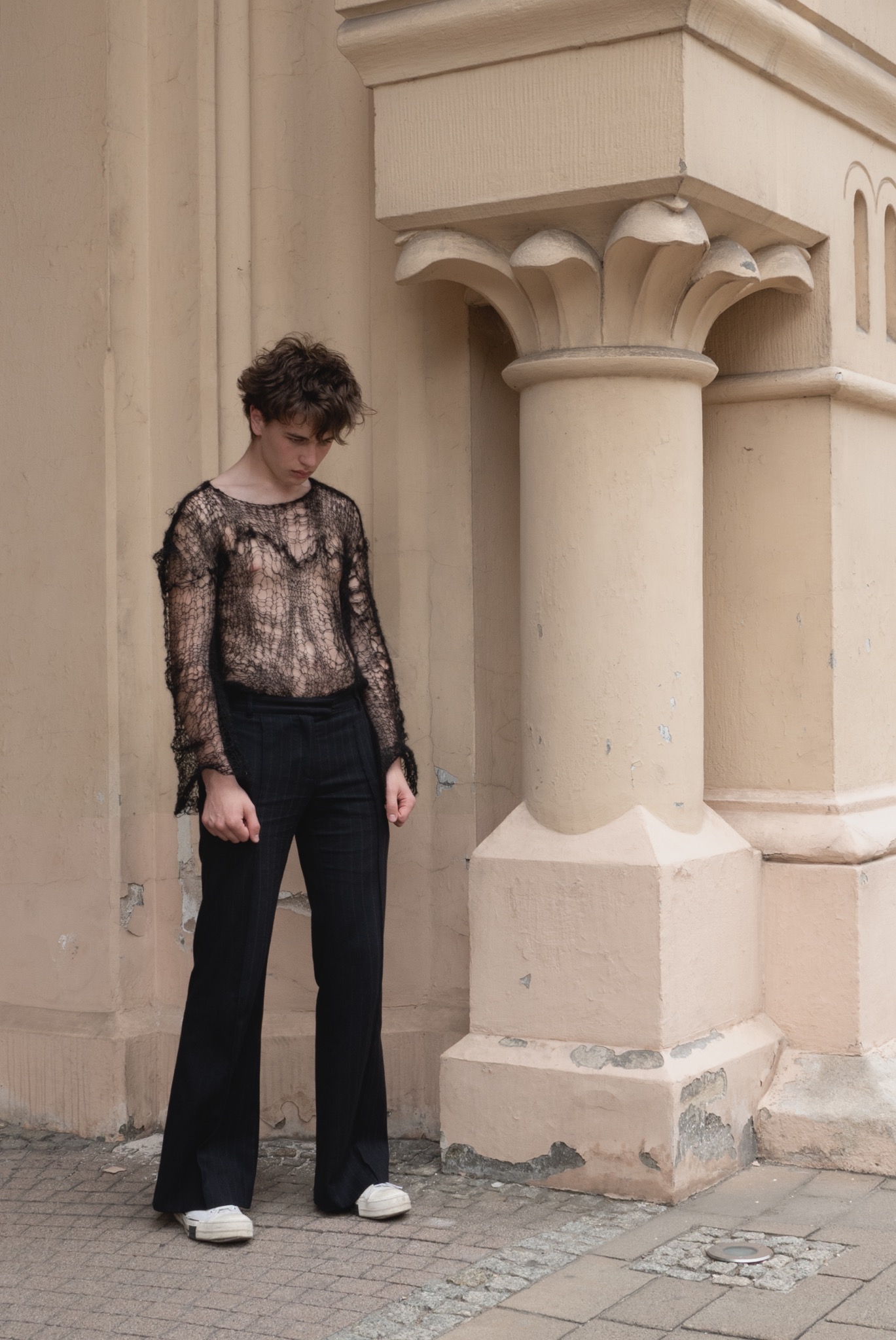 Młody mężczyzna w ażurowej bluzce i szerokich spodniach pozuje przy budynku z kolumnami, spoglądając w dół.