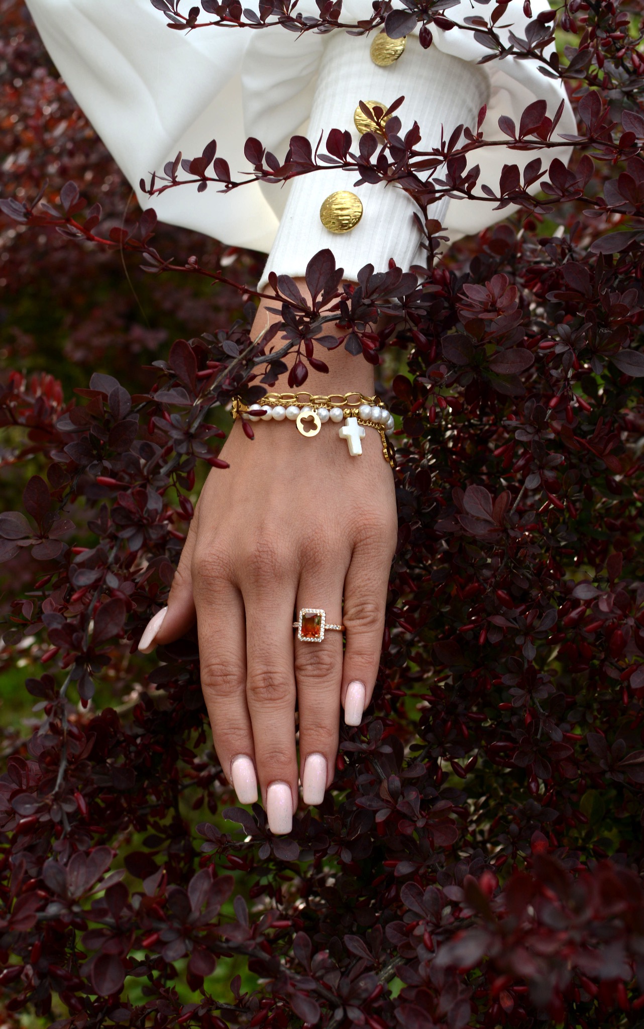 Dłoń kobiety z jasnym manicurem i złotym pierścionkiem z pomarańczowym kamieniem, ozdobiona bransoletkami z pereł i złotych łańcuszków z zawieszkami, na tle bordowych liści krzewu i białej koszuli...