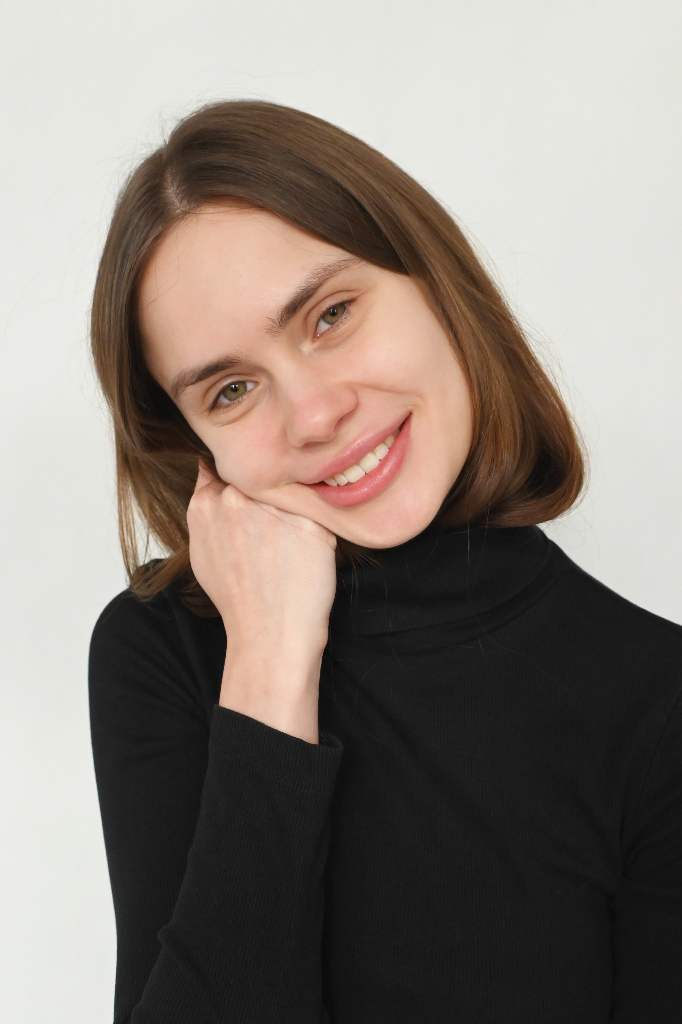 Portret młodej kobiety z brązowymi włosami, uśmiechającej się i opierającej policzek na dłoni, ubrana w czarny golf na białym tle.