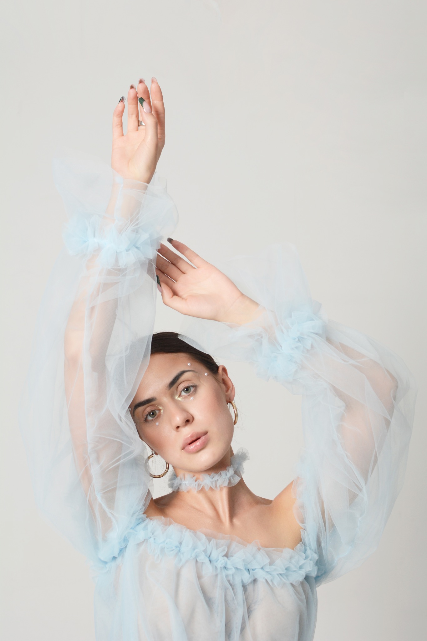 Portret kobiety w błękitnej, tiulowej sukience z bufiastymi rękawami i chokerem, z delikatnym makijażem i złotymi kolczykami kołami, pozującej z rękami uniesionymi do góry.