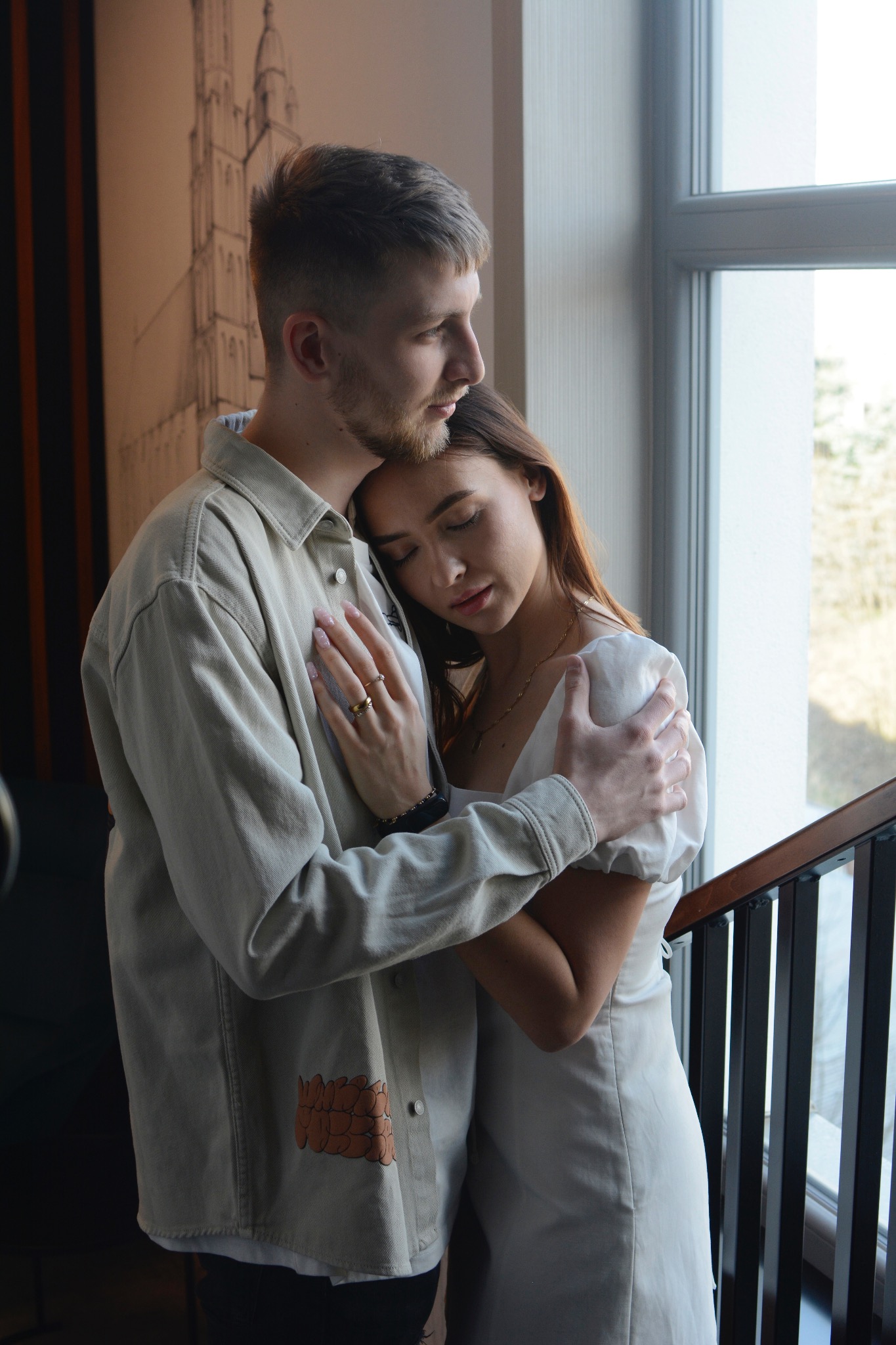 Para zakochanych obejmuje się blisko okna: mężczyzna w dżinsowej kurtce z aplikacją, kobieta w białej sukience z zamkniętymi oczami, oboje w czułym uścisku.