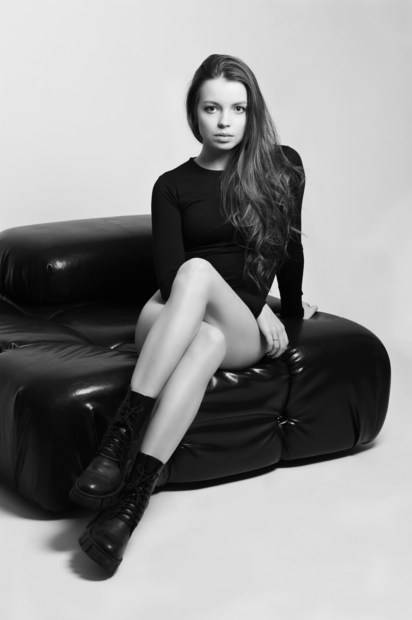 Portret kobiety w czarnym body i botkach, siedzącej na skórzanej pufie w studio, zdjęcie czarno-białe.