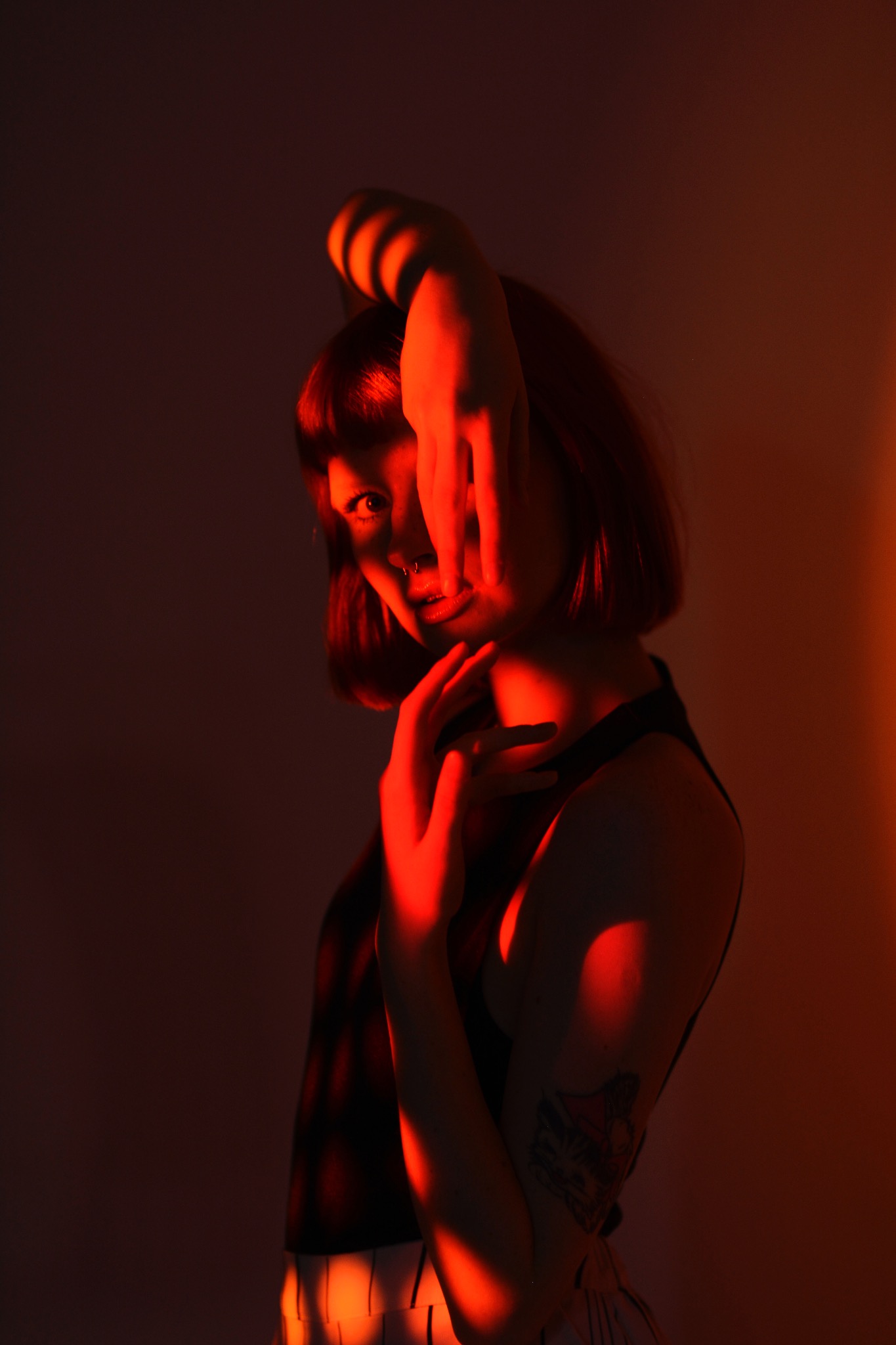 Portret kobiety z piercingiem w nosie, zasłaniającej twarz rękami, oświetlonej czerwonym światłem, z widocznym tatuażem na ramieniu.