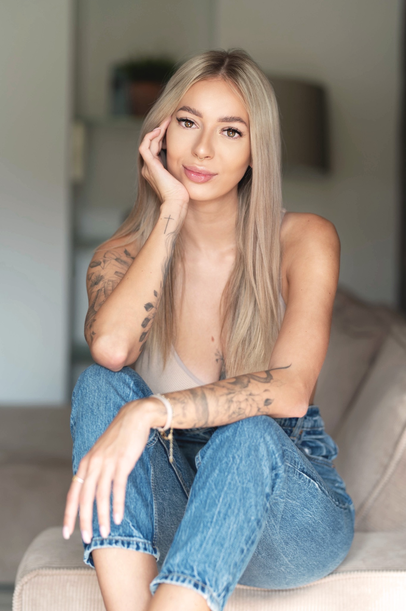 Portret młodej kobiety z tatuażami na ramionach, siedzącej na beżowej sofie w dżinsach i topie, z ręką podpartą pod brodą i delikatnym uśmiechem.