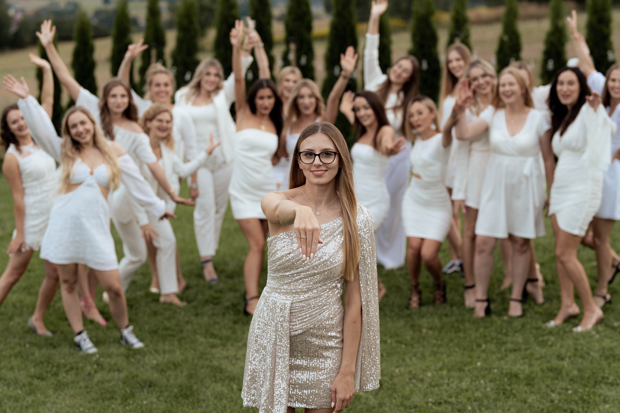 Panna młoda w błyszczącej sukience prezentuje pierścionek zaręczynowy na tle druhen w białych sukienkach, podczas sesji plenerowej z zielenią w tle.