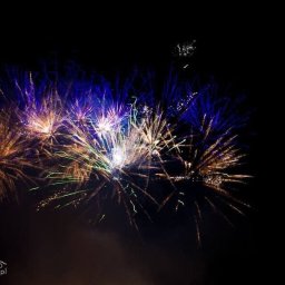 Sultex Beata Sulis - Wybuchające na nocnym niebie niebiesko-złote fajerwerki z zielonymi smugami, uchwycone w kadrze z lekkim efektem rozmycia ruchu.