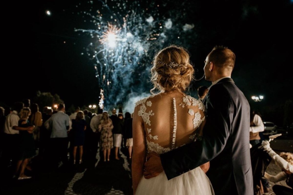 Para młoda wpatruje się w fajerwerki na tle nocnego nieba, otoczona gośćmi weselnymi.