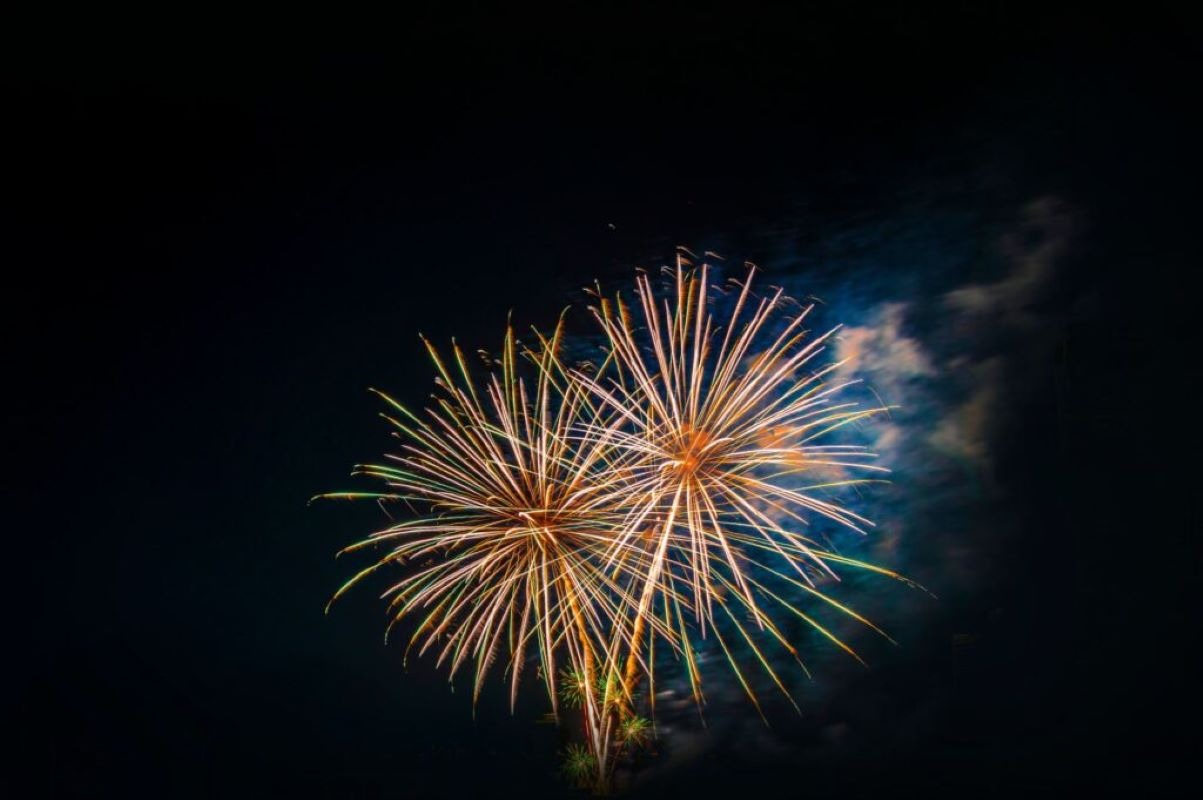 Dwa wybuchy fajerwerków na nocnym niebie, z widocznym dymem i złotymi, zielonymi i białymi promieniami.