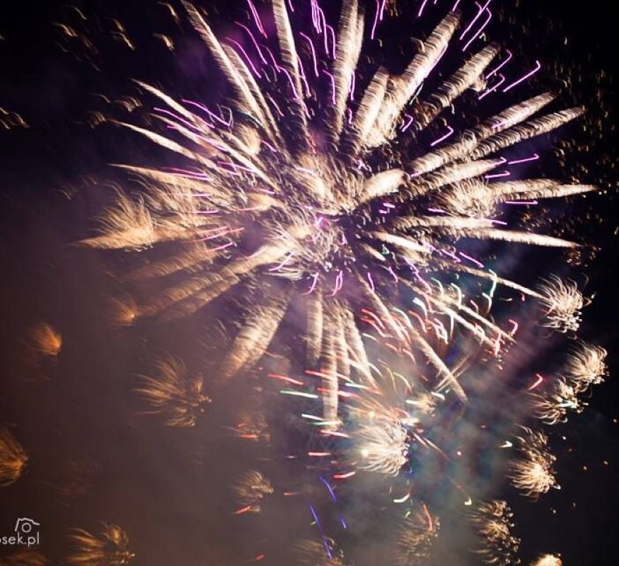 Wybuchający fajerwerk na nocnym niebie z długimi smugami światła w kolorach białym, fioletowym, czerwonym i zielonym, otoczony dymem.