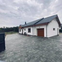 Nowoczesny dom jednorodzinny z szarą kostką brukową na podjeździe, widoczna brama wjazdowa i garażowa, pochmurne niebo.