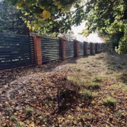 Długi, nowoczesny płot z poziomych, ciemnych desek i ceglanych słupk&oacute;w, widoczny z perspektywy ziemi pokrytej opadłymi liśćmi, częściowo zacieniony przez drzewa.