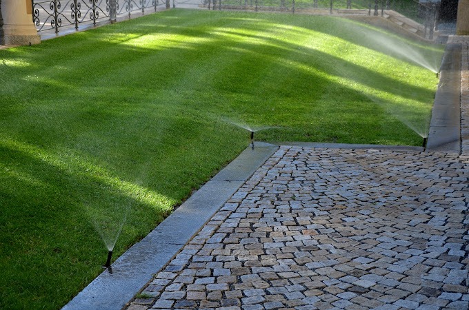 Automatyczne nawadnianie trawnika z kostką brukową w tle, widoczne zraszacze w akcji.