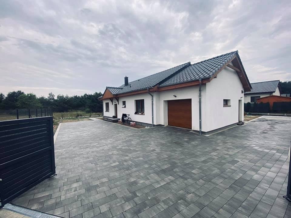 Nowoczesny dom jednorodzinny z szarą kostką brukową na podjeździe, widoczna brama wjazdowa i garażowa, pochmurne niebo.