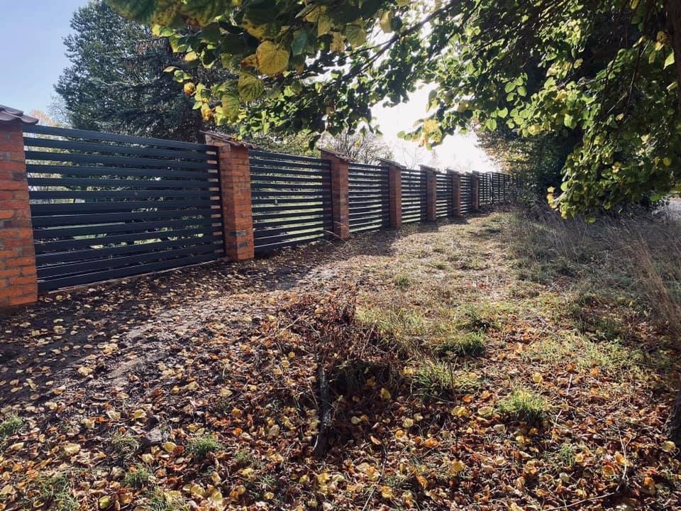 Długi, nowoczesny płot z poziomych, ciemnych desek i ceglanych słupków, widoczny z perspektywy ziemi pokrytej opadłymi liśćmi, częściowo zacieniony przez drzewa.