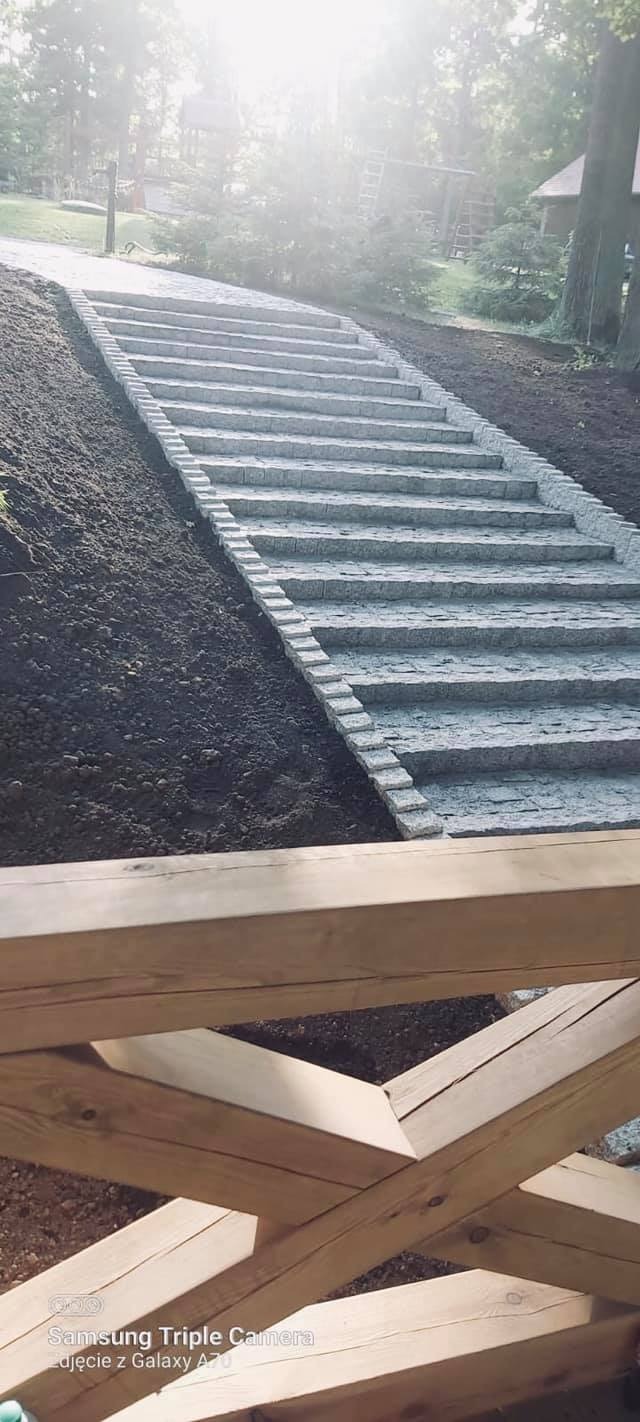Kamienne schody prowadzące w górę wzgórza, z widoczną konstrukcją drewnianą w dolnej części kadru i drzewami w tle.