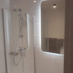 OGULBUD - Nowoczesna kabina prysznicowa z szklanymi drzwiami i chromowaną armaturą, widoczne białe płytki ceramiczne i podświetlane lustro z zaokrąglonymi rogami odbijające fragment toalety.