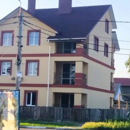 OGULBUD - Trzypiętrowy dom z beżową elewacją i bordowymi akcentami, widoczne balkony z kutymi balustradami, ciemny dach, drewniany płot oraz słup energetyczny z ogłoszeniem.