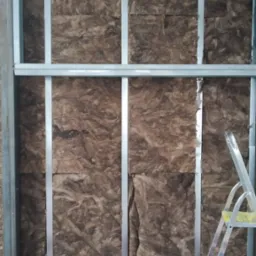 Wnętrze ściany z izolacją z wełny mineralnej pomiędzy metalowymi profilami, widoczny fragment rury wentylacyjnej, drabina i narzędzia na pierwszym planie.