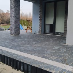 OGULBUD - Wykończony taras z szarej kostki brukowej ułożonej w jodełkę, z jasnym pasem oddzielającym od ziemi, filar z kamienia dekoracyjnego, widoczne elementy elewacji domu i osoba w tle.