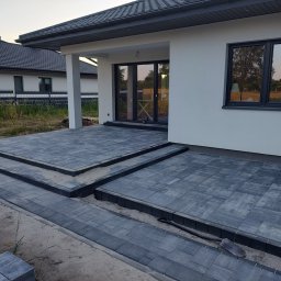 OGULBUD - Nowoczesny taras z szarej kostki brukowej przed domem z białymi ścianami i ciemnymi oknami, widoczne schody tarasowe i przygotowane podłoże z piasku.