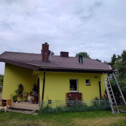 OGULBUD - Dwaj mężczyźni pracują na brązowym dachu żółtego domu jednorodzinnego, jeden przy kominie, drugi przy krawędzi, z drabiną opartą o ścianę i osobą z psem na ganku.