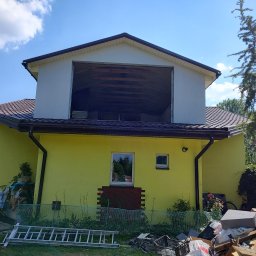 OGULBUD - Dwukondygnacyjny dom z żółtą elewacją i otwartym poddaszem, widoczne materiały budowlane i drabina na trawniku, sugerujące prace remontowe.