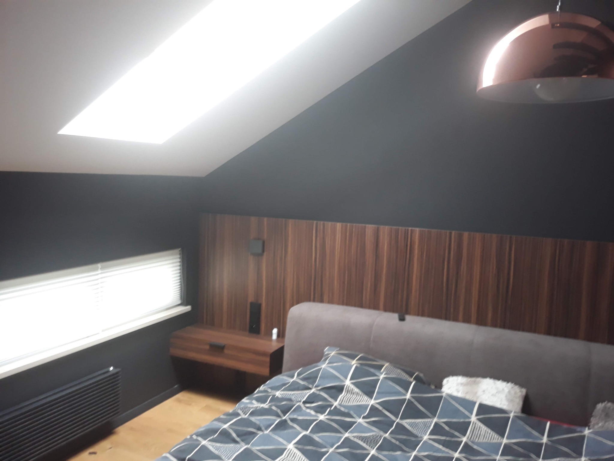 Sypialnia na poddaszu z ciemnymi ścianami, drewnianym panelem za łóżkiem i miedzianą lampą wiszącą.