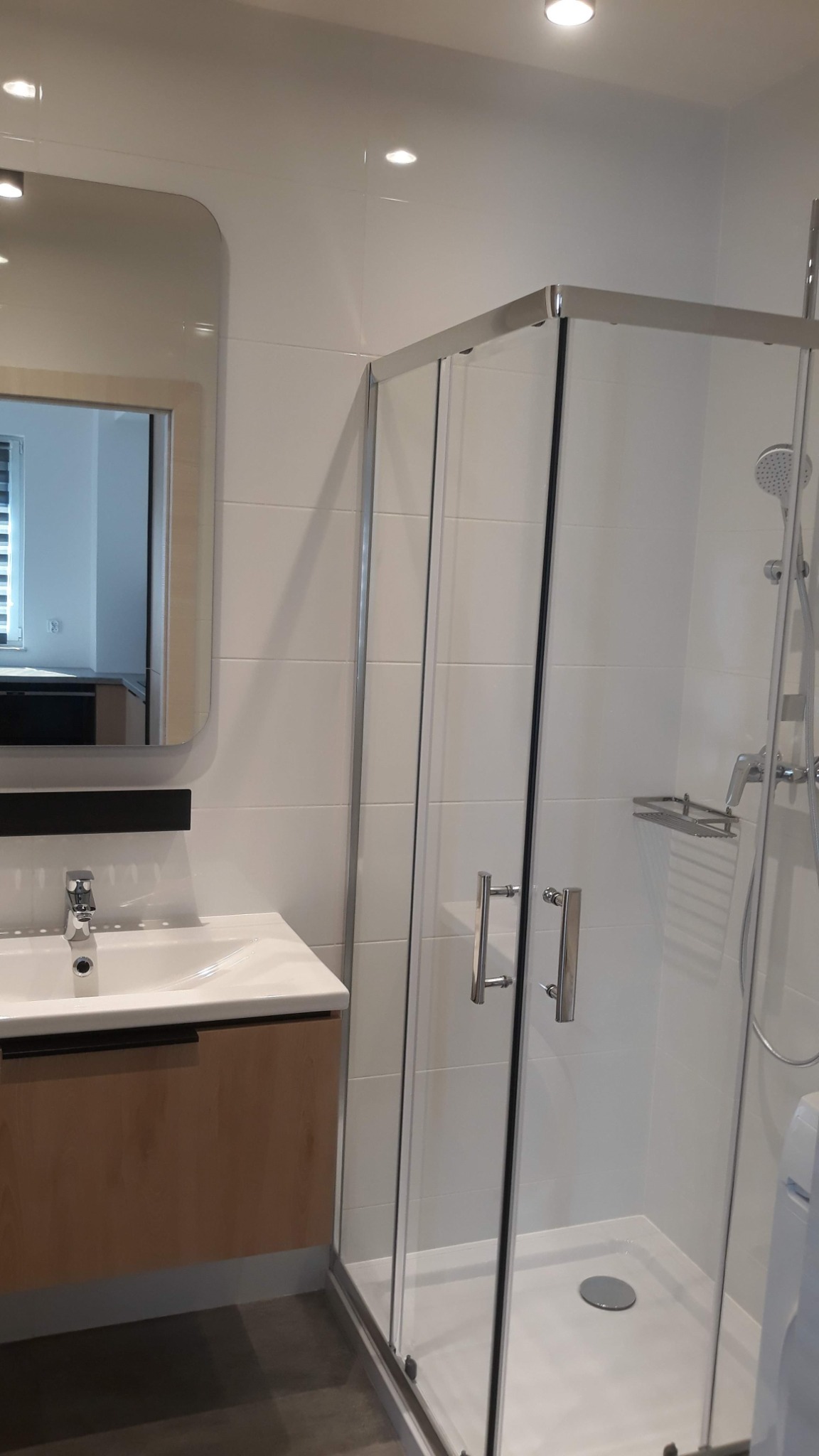 Nowoczesna łazienka z kabiną prysznicową z chromowanymi profilami, umywalką z szafką w kolorze jasnego drewna i dużym lustrem z zaokrąglonymi rogami, oświetlona punktowymi lampami sufitowymi.