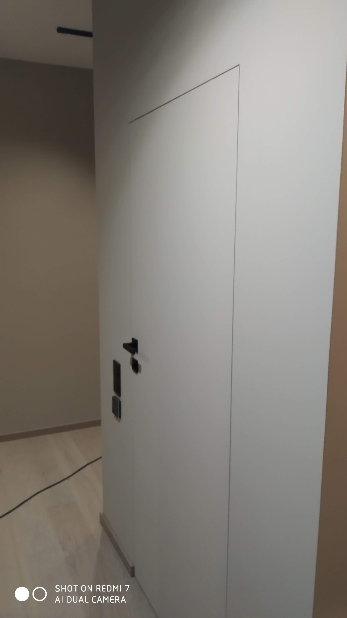 Białe, ukryte drzwi w ścianie, z czarną klamką i panelem sterowania, z subtelną linią oddzielającą od ściany, na tle jasnej podłogi i ściany w odcieniach beżu.