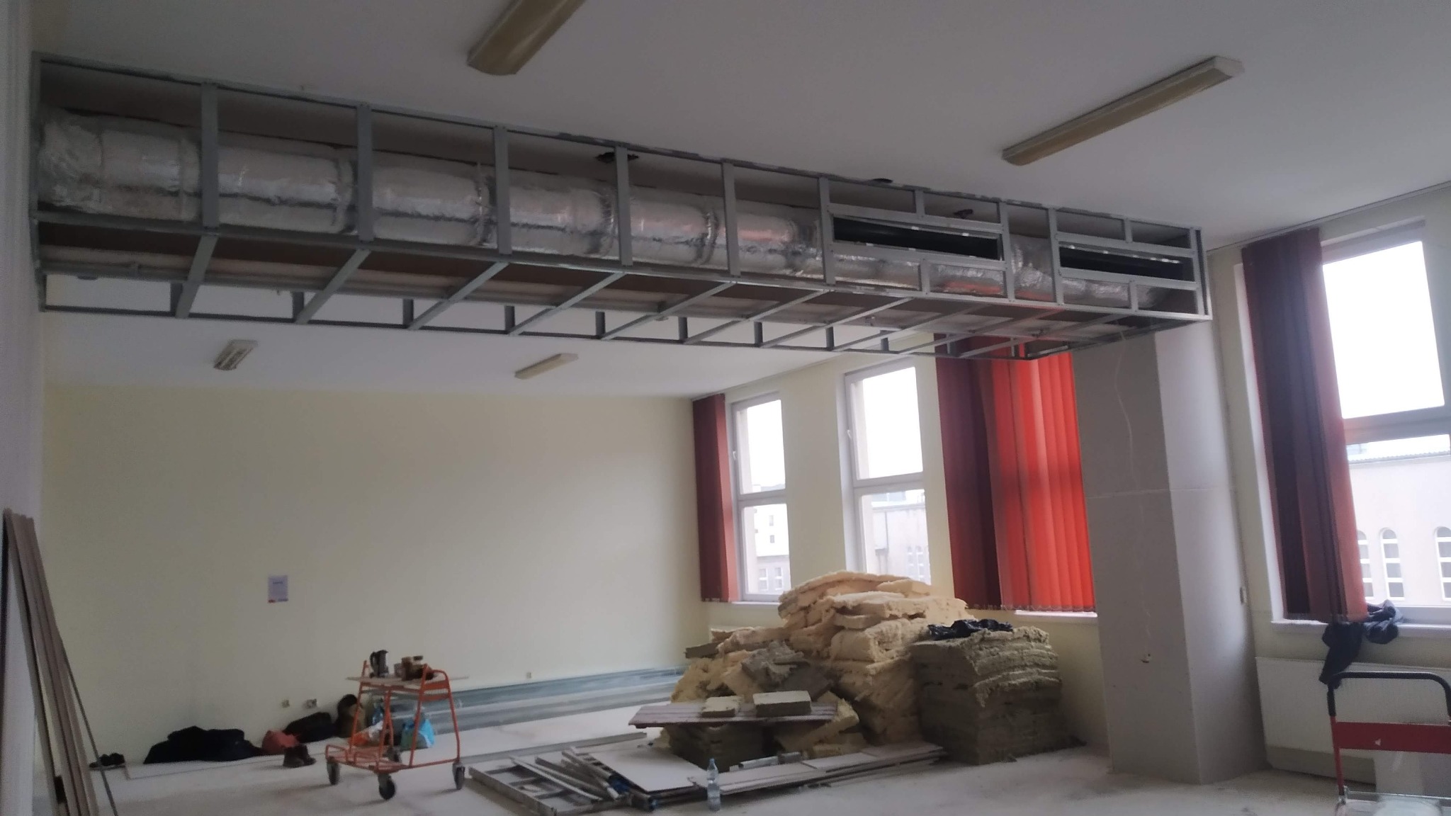 Wnętrze w trakcie prac: metalowa konstrukcja podwieszanego sufitu z izolacją, stos wełny mineralnej na podłodze, widoczne okna z czerwonymi zasłonami, wózek z narzędziami.