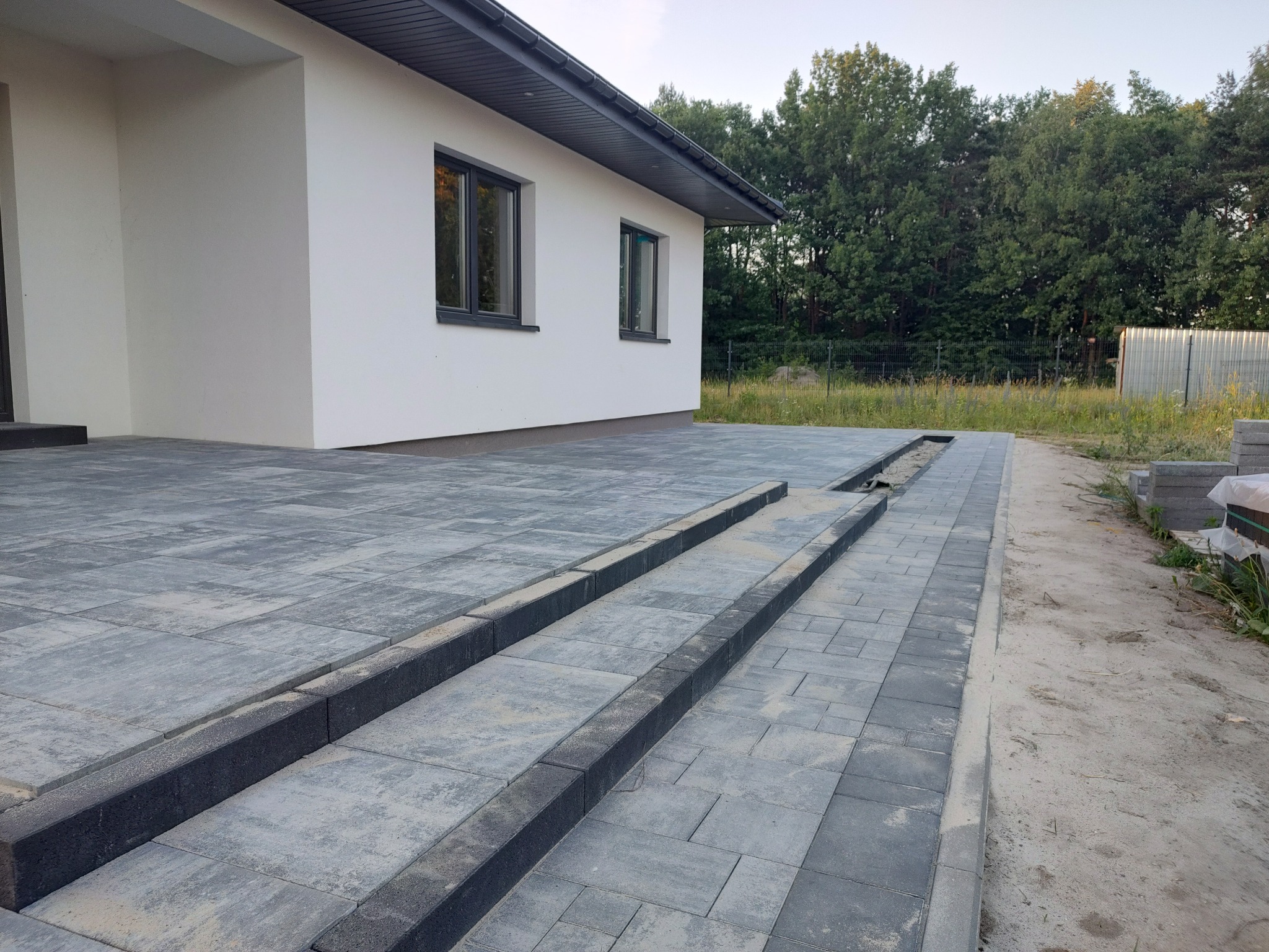 Ułożona kostka brukowa w odcieniach szarości przed nowoczesnym domem z białymi ścianami i ciemnymi oknami, widoczne obrzeża i schody.