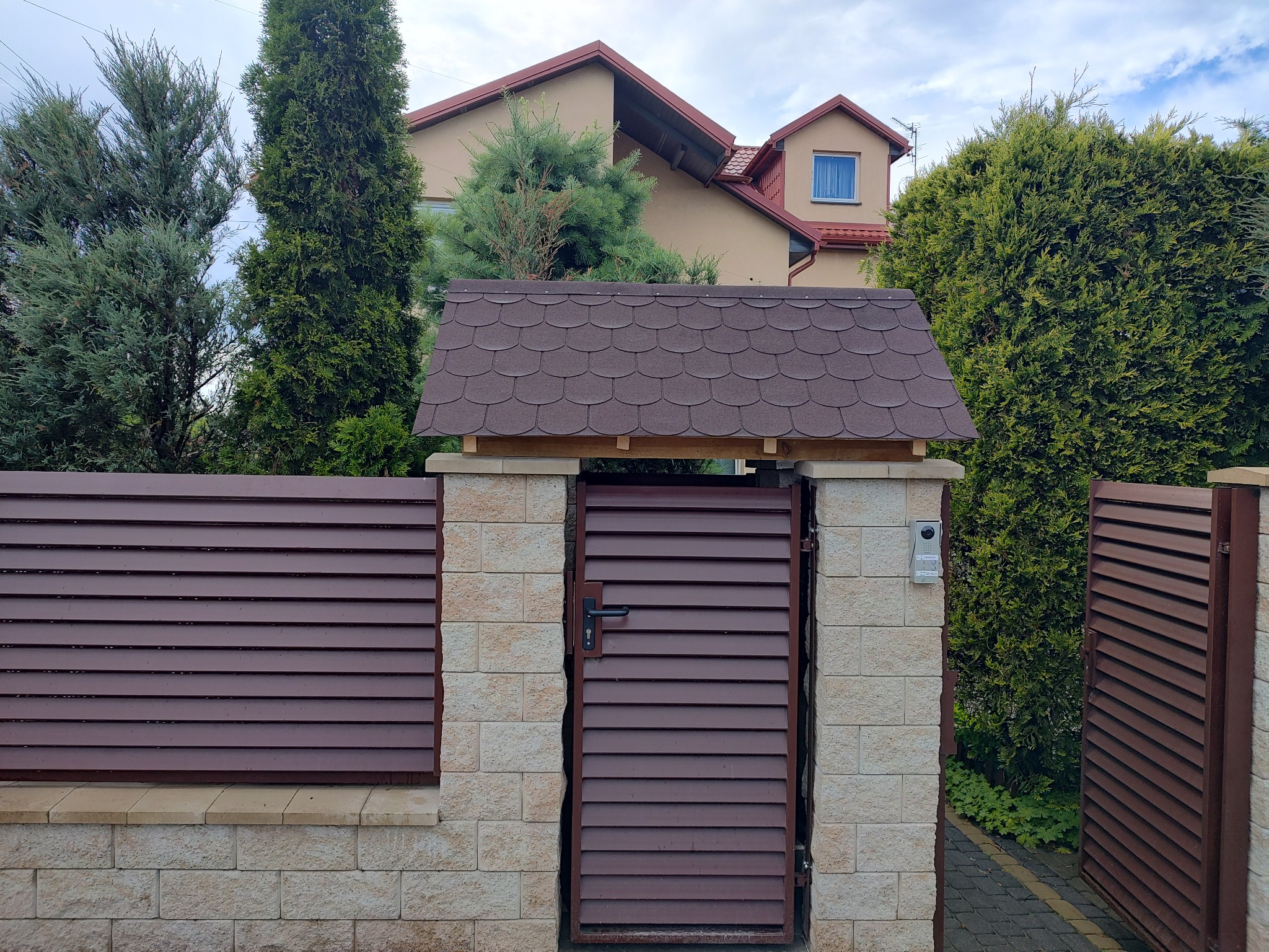 Brązowa, pozioma brama lamelowa z daszkiem z brązowej dachówki bitumicznej, wkomponowana w ogrodzenie z jasnych, ciosanych kamieni, na tle domu z beżową elewacją i zielonych drzew.