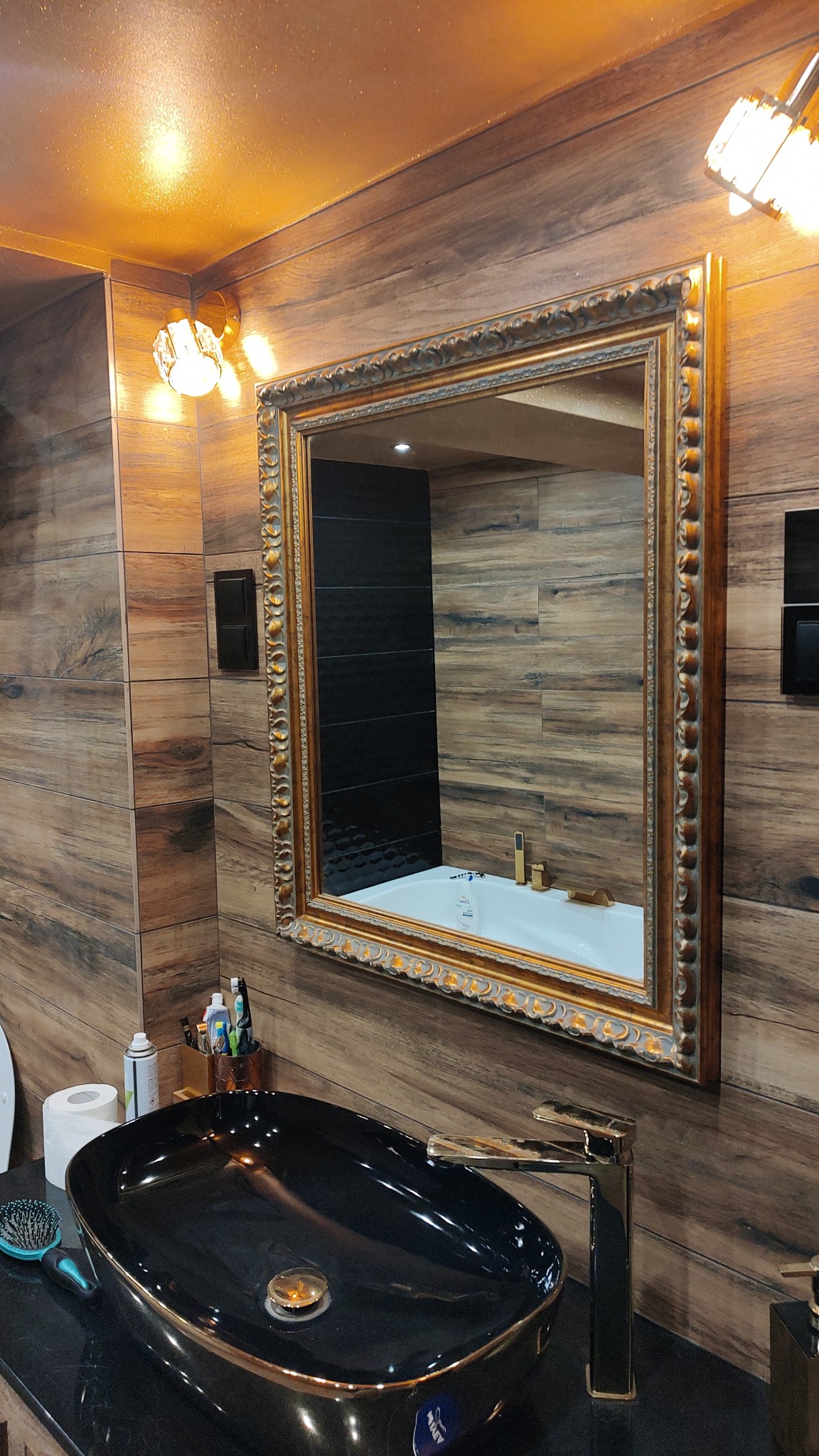 Nowoczesna łazienka z czarną, owalną umywalką nablatową z złotymi akcentami, lustrem w ozdobnej, złotej ramie i drewnopodobnymi płytkami na ścianach.