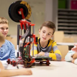 Grupa dzieci obserwuje działanie drukarki 3D w klasie podczas warsztatów edukacyjnych.