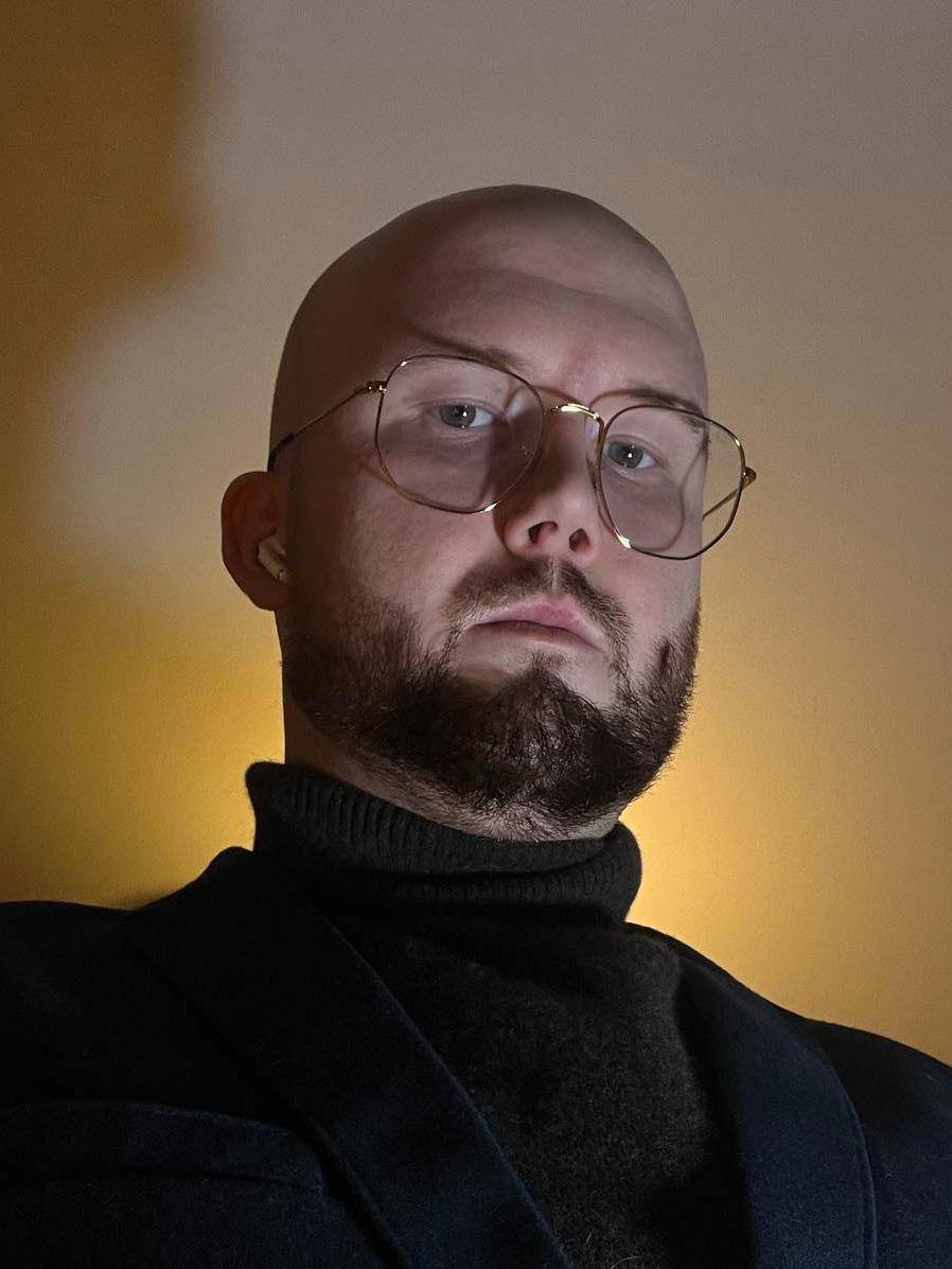 Portret mężczyzny z brodą w okularach, ubrany w ciemny golf i marynarkę, oświetlony od dołu ciepłym światłem.