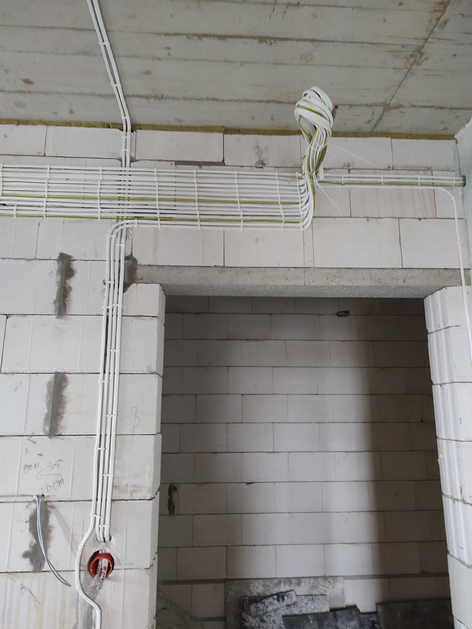 Białe kable elektryczne ułożone w peszlach na ścianie z białych bloczków, widoczne otwory pod gniazdka, surowy stan wykończenia wnętrza.