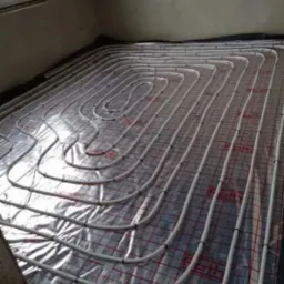 Białe rury ogrzewania podłogowego ułożone spiralnie na folii z siatką w nowym budynku, widoczne czarne mocowania rur.