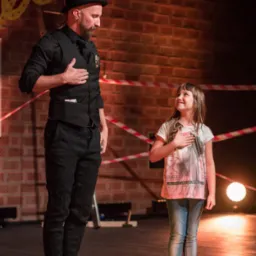Mężczyzna w czarnym stroju i kapeluszu oraz dziewczynka na scenie, oboje z rękami na sercu, występ na tle ceglanej ściany z dekoracjami i światłami.