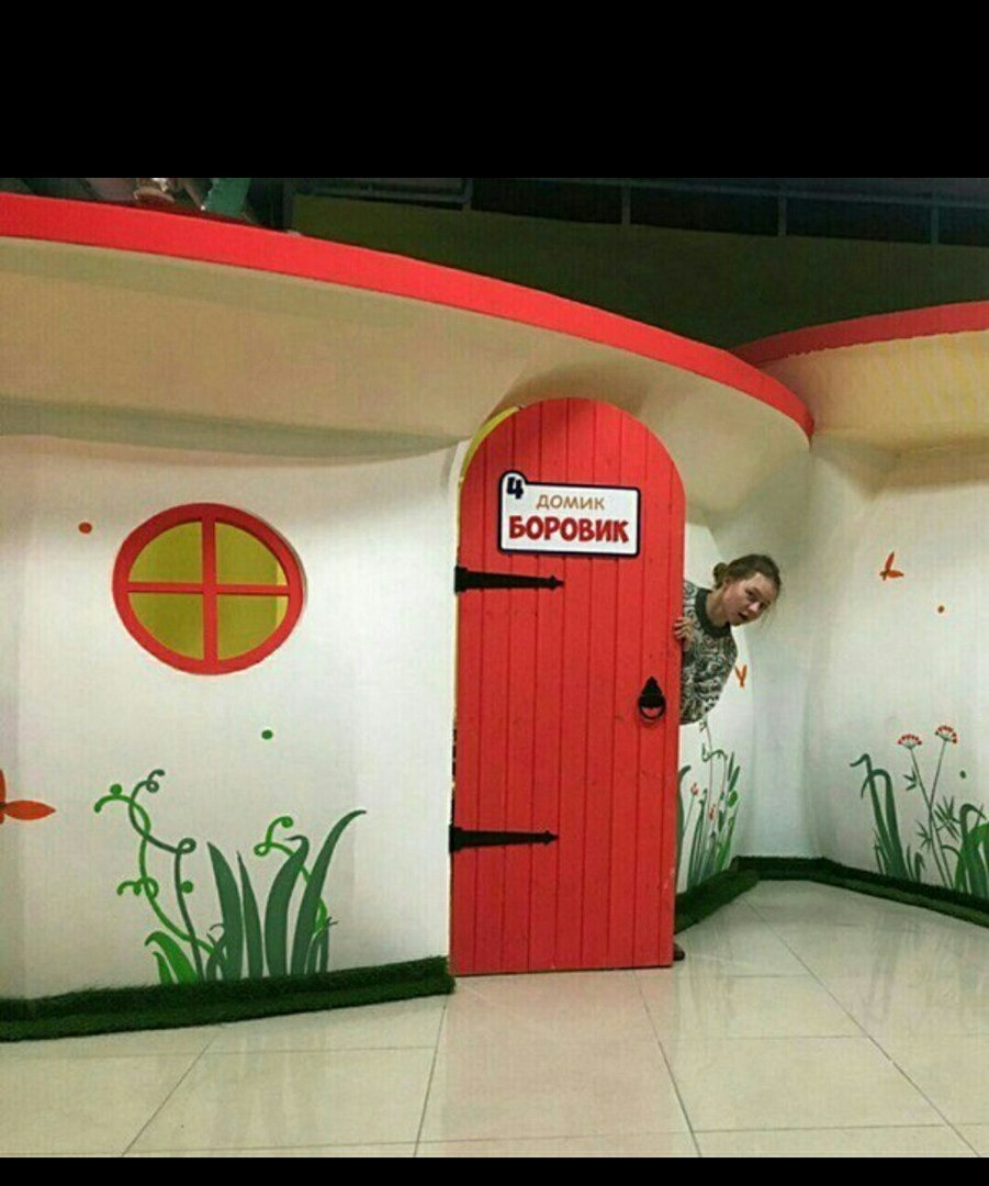 Wnętrze pokoju zabaw w kształcie domku z czerwoną okrągłą oknem i drzwiami, na ścianach zielone malowidła roślinne, a zza drzwi wygląda uśmiechnięta dziewczyna.