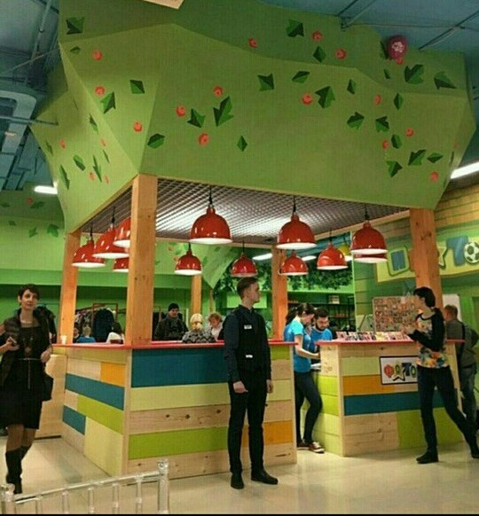 Wnętrze sali zabaw dla dzieci z ladą recepcyjną w kształcie drzewa z czerwonymi lampami, obsługa i klienci w tle.