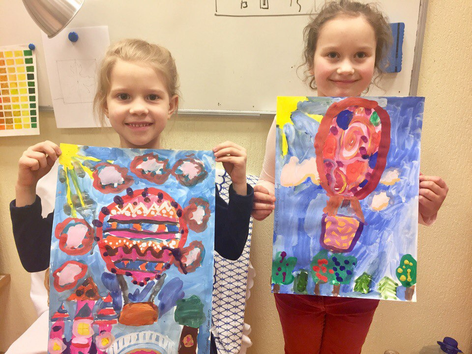 Dwie uśmiechnięte dziewczynki prezentują swoje prace plastyczne: akwarelowe obrazy przedstawiające balony na tle nieba i krajobrazu, z widocznymi śladami pędzla i dziecięcą ekspresją.