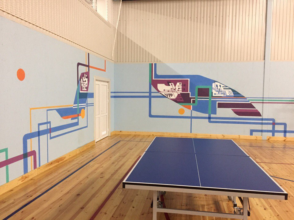 Wnętrze sali gimnastycznej z abstrakcyjnym, geometrycznym malunkiem ściennym w odcieniach niebieskiego, pomarańczowego, fioletowego i zielonego, uzupełnionym o motywy przypominające schematy...