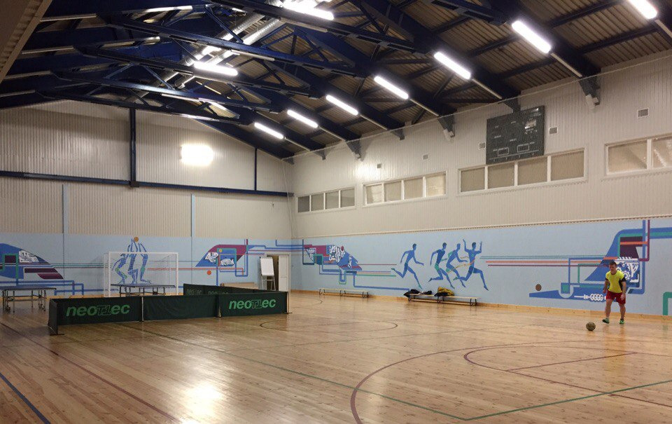 Wnętrze hali sportowej z drewnianą podłogą, pomalowaną ścianą z motywami sportowymi i grającym chłopcem w prawym dolnym rogu.