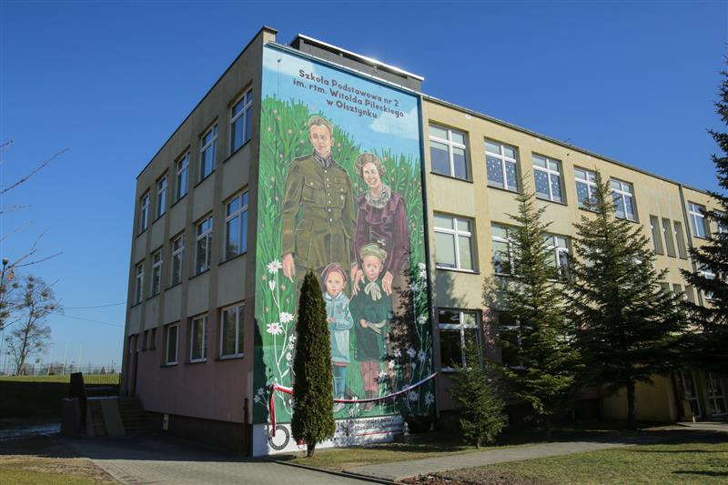 Mural na ścianie budynku szkoły podstawowej przedstawiający portret rodziny Witolda Pileckiego na tle łąki z kwiatami.